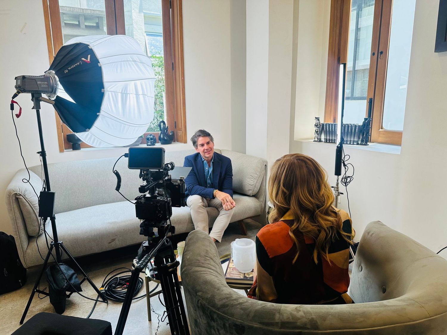 Videographer films interview between two people seated in bright naturally lit living room.