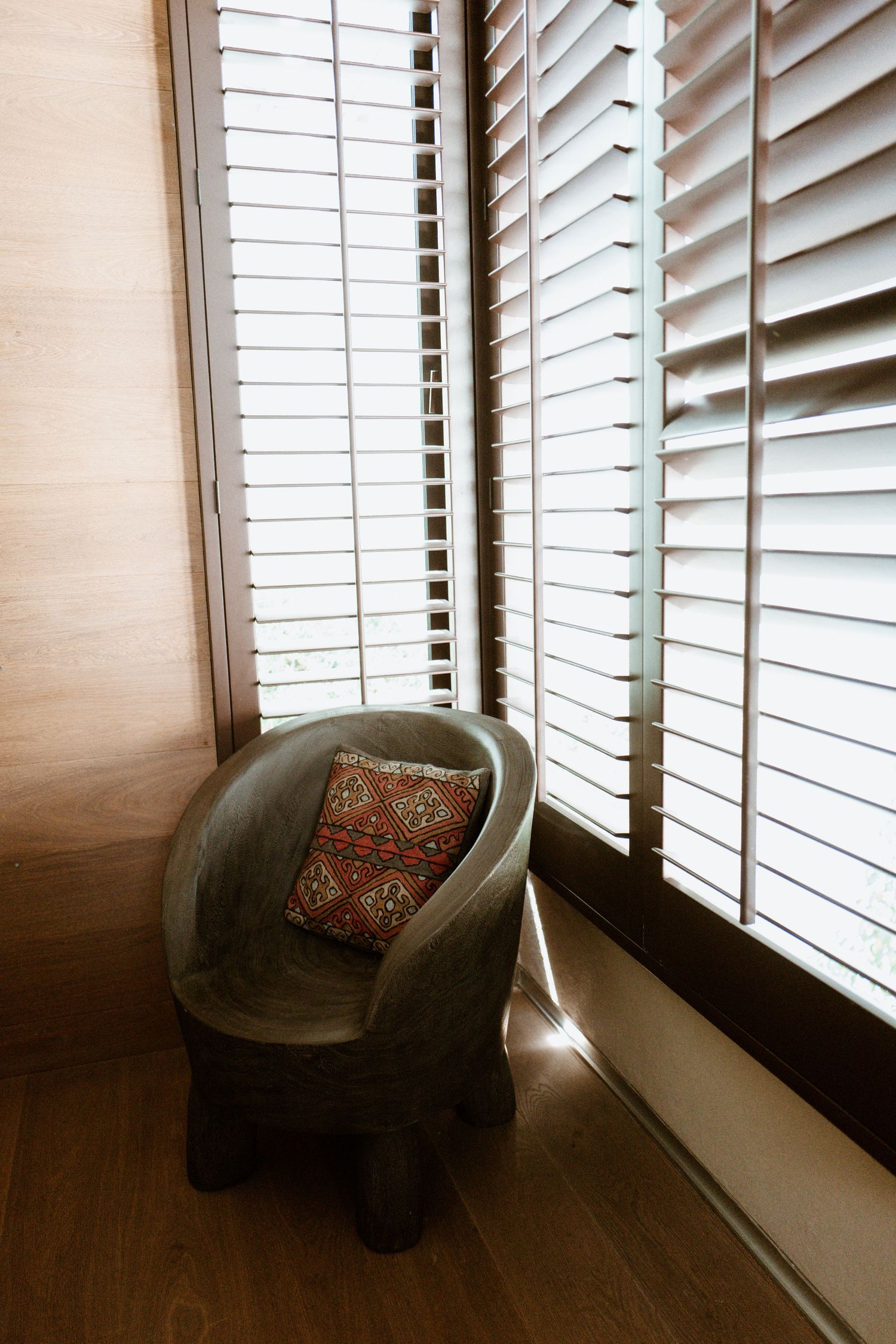 Gray barrel chair with red patterned pillow beside wooden shuttered windows on hardwood floor.