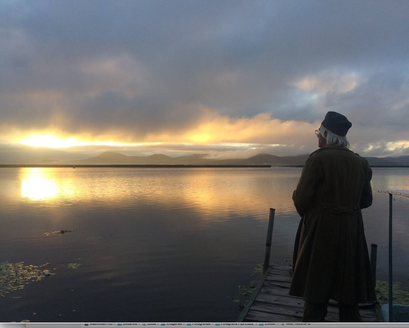 Person in long coat standing on dock watching golden sunset over calm lake.