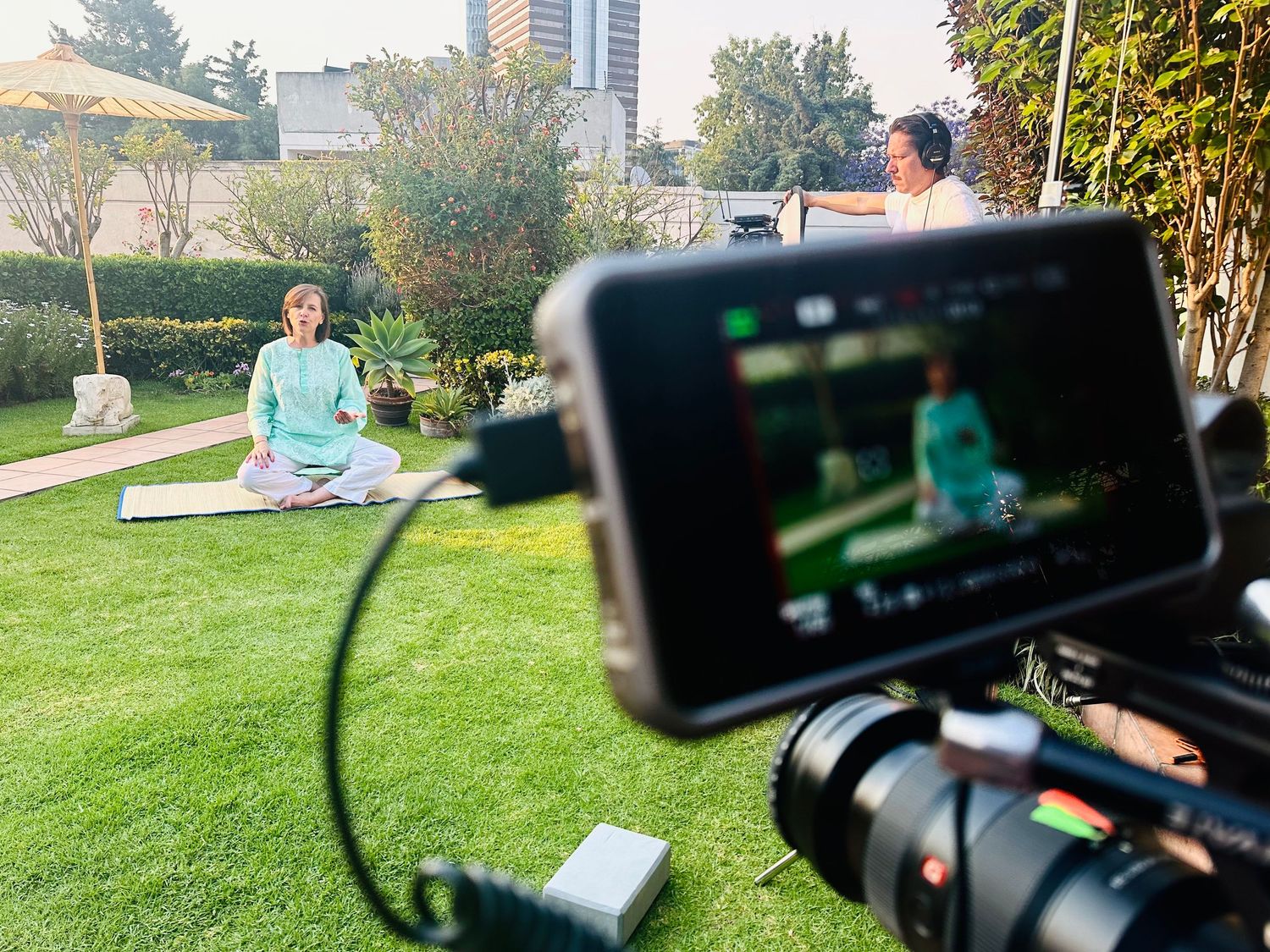 Outdoor video production filming a woman meditating in lotus pose on green lawn in daylight.