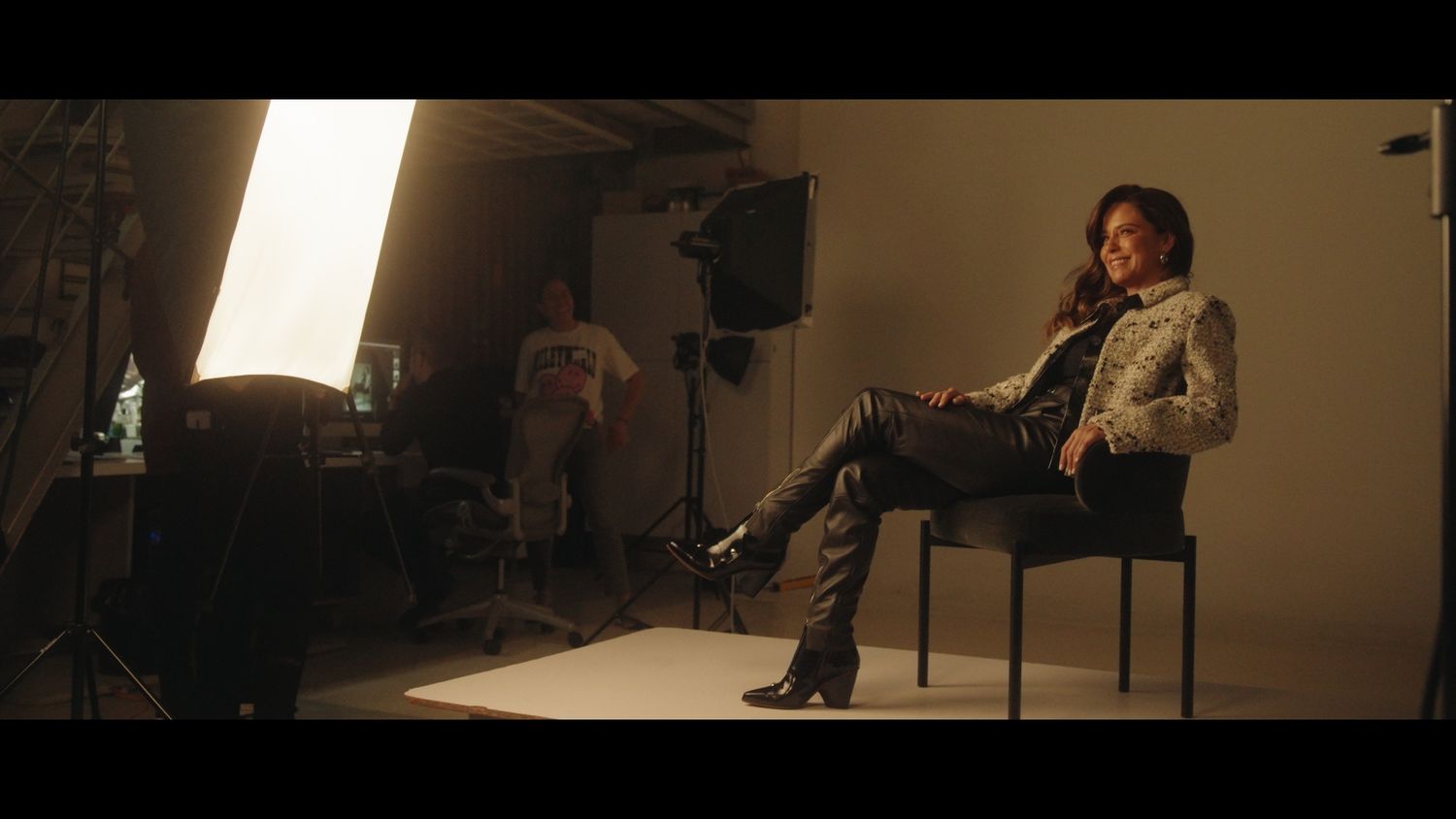 Female model seated on chair during professional photo shoot with softbox lighting in a studio setting.
