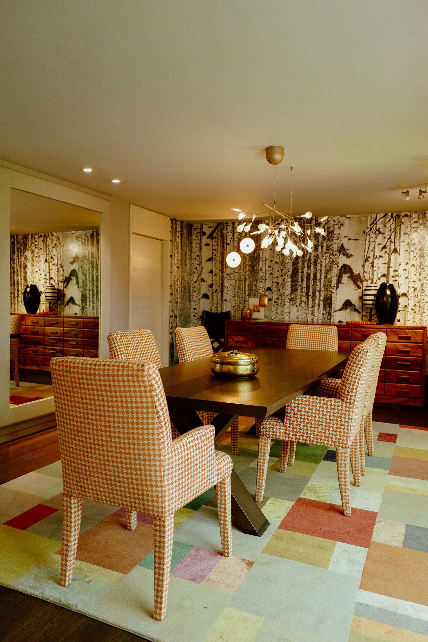 Stylish dining area featuring houndstooth upholstered chairs, colorful patchwork rug, and birch wallpaper.