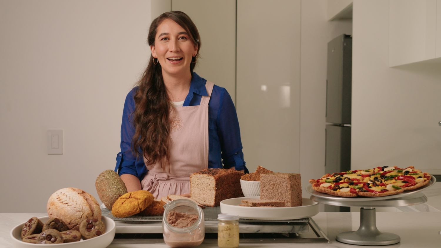 Smiling woman in apron standing behind a kitchen counter with various baked goods.