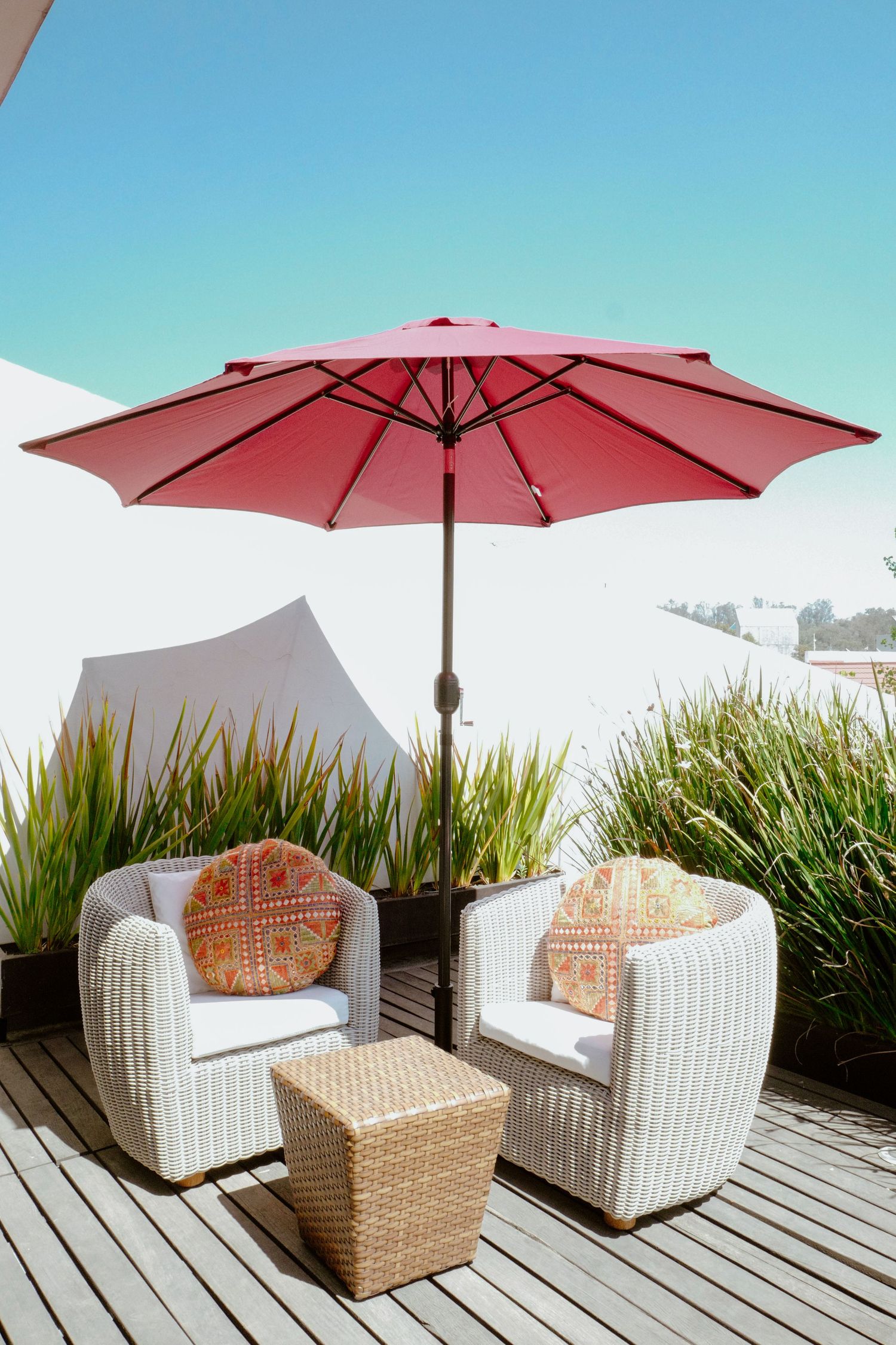 White wicker chairs with orange pillows under a red patio umbrella on a wooden deck.