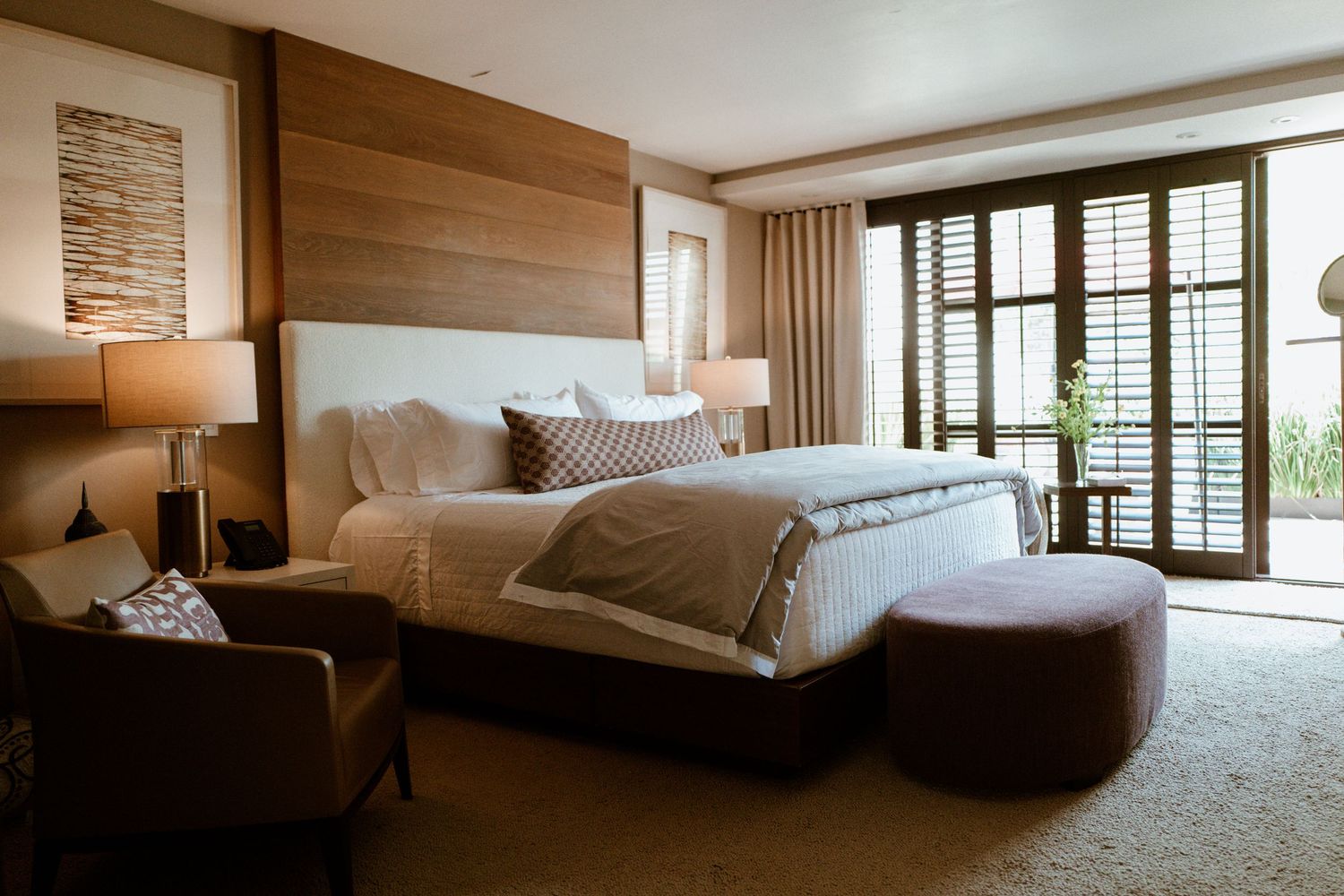 Elegant hotel bedroom with king bed, wood accent wall, shuttered doors, and warm lighting.