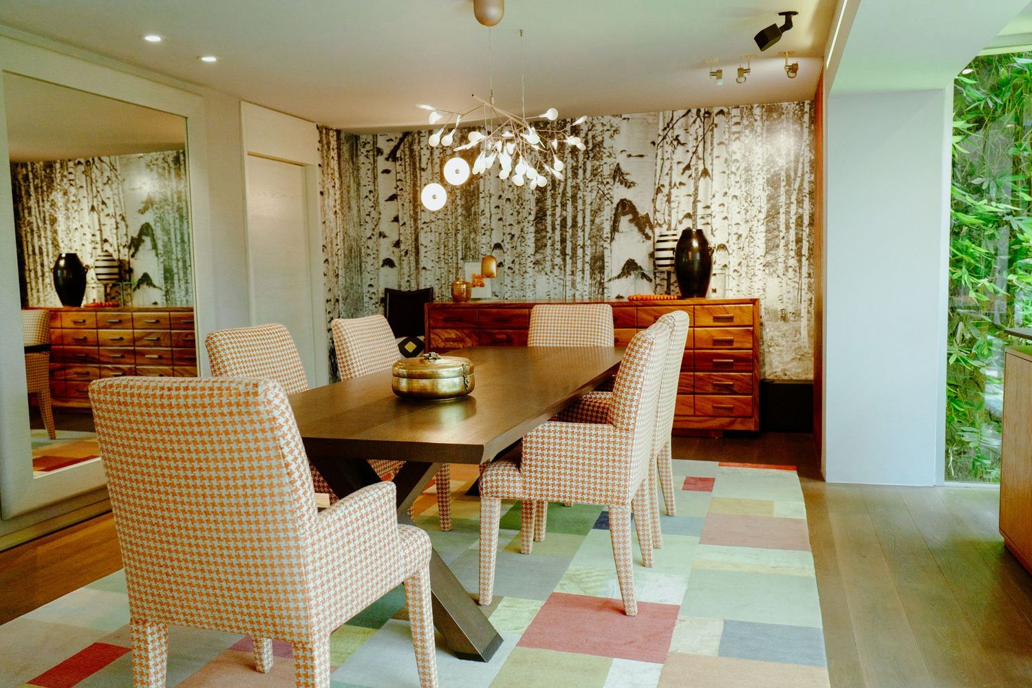 Elegant dining room with birch tree wallpaper, houndstooth chairs, wooden sideboard, and modern chandelier.