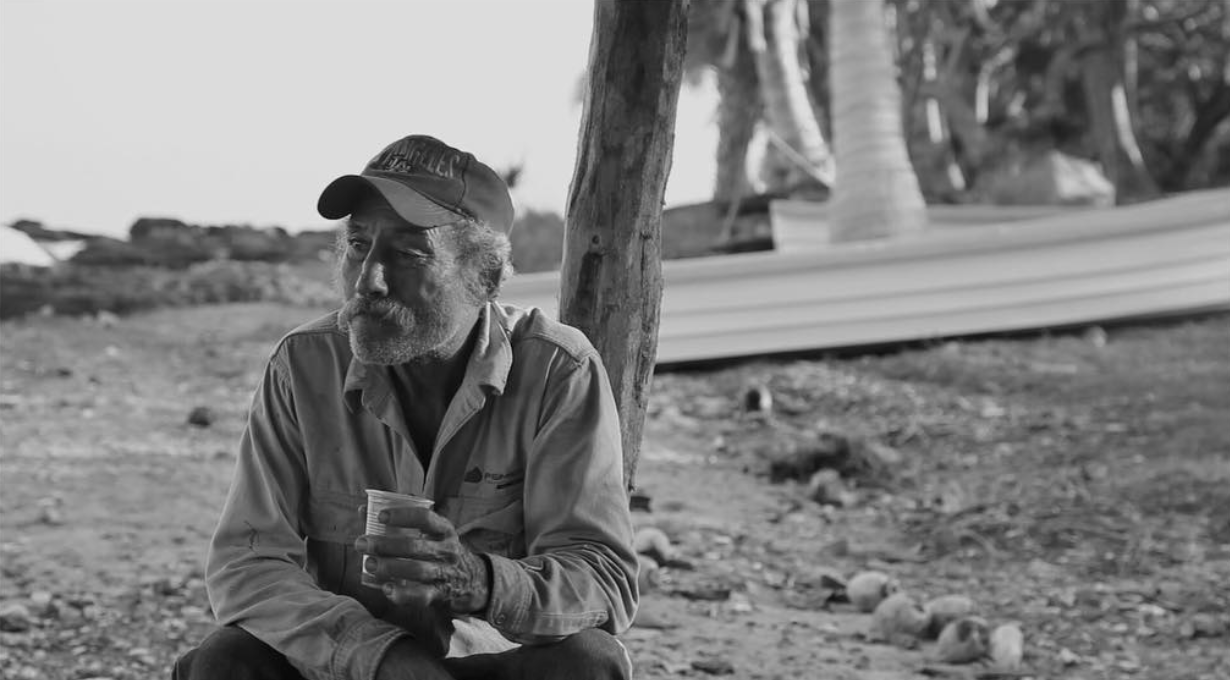 Elderly man in cap sits outdoors holding a cup, black and white photo with trees in background.