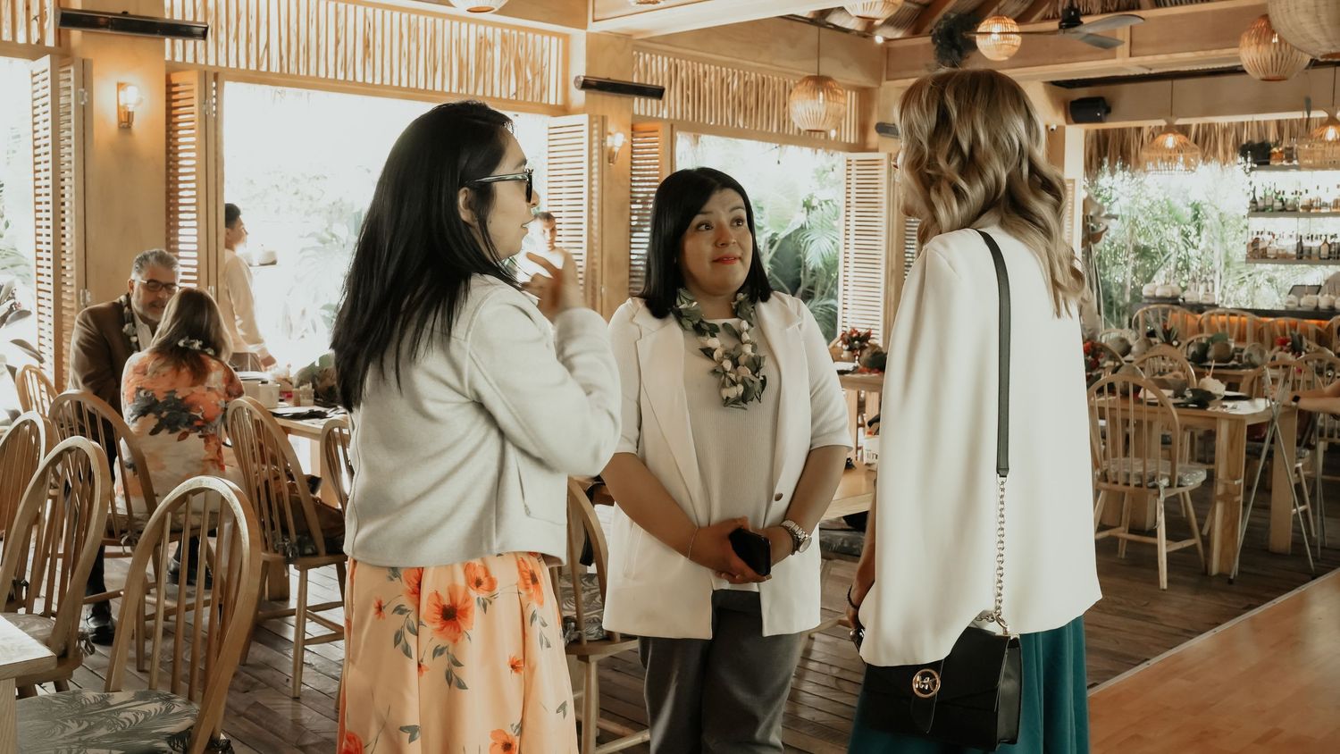 Three women in white tops having a conversation inside a bright tropical-themed restaurant setting.
