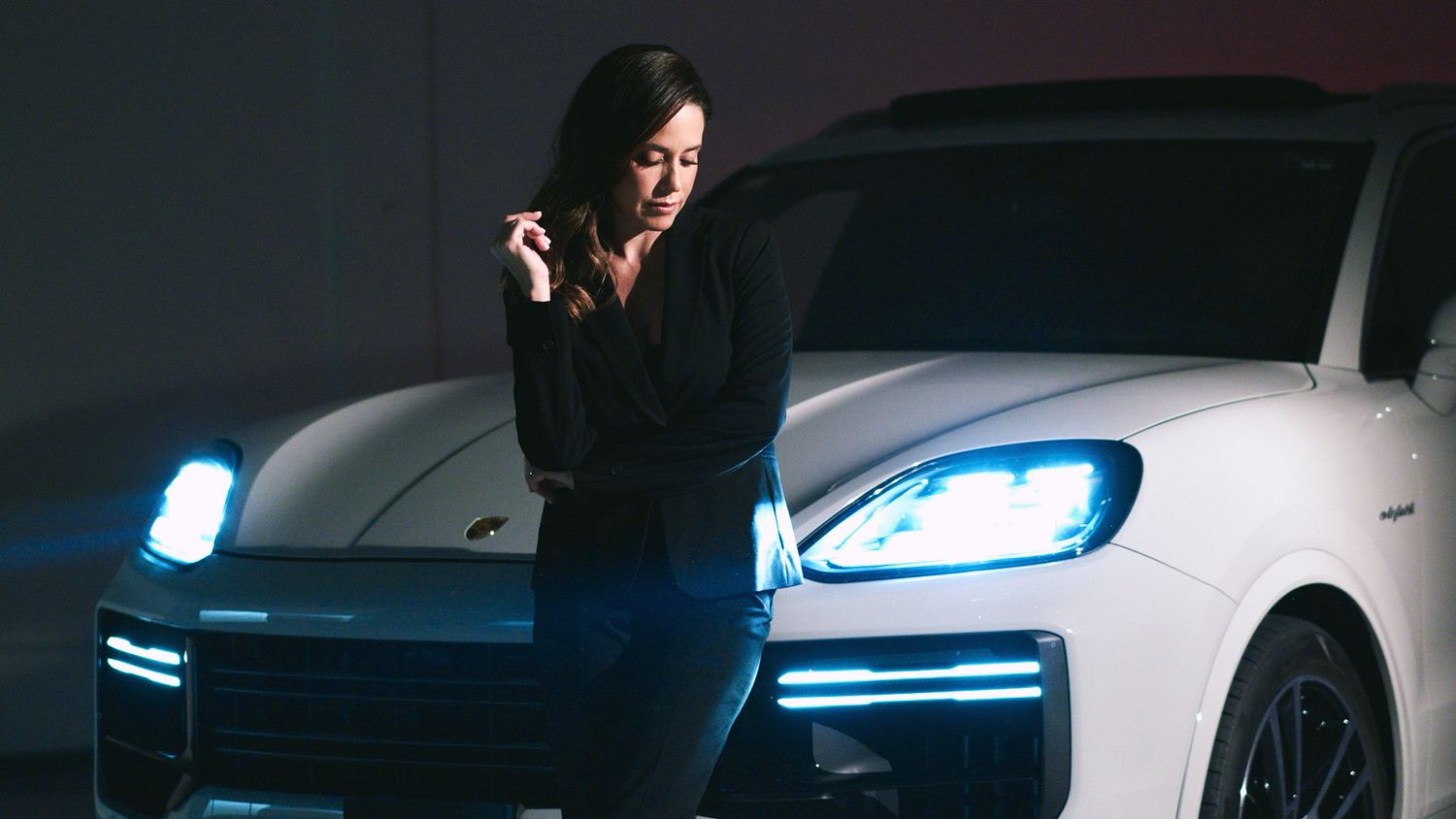 Woman in black outfit posing beside white Porsche Cayenne with glowing headlights at night.