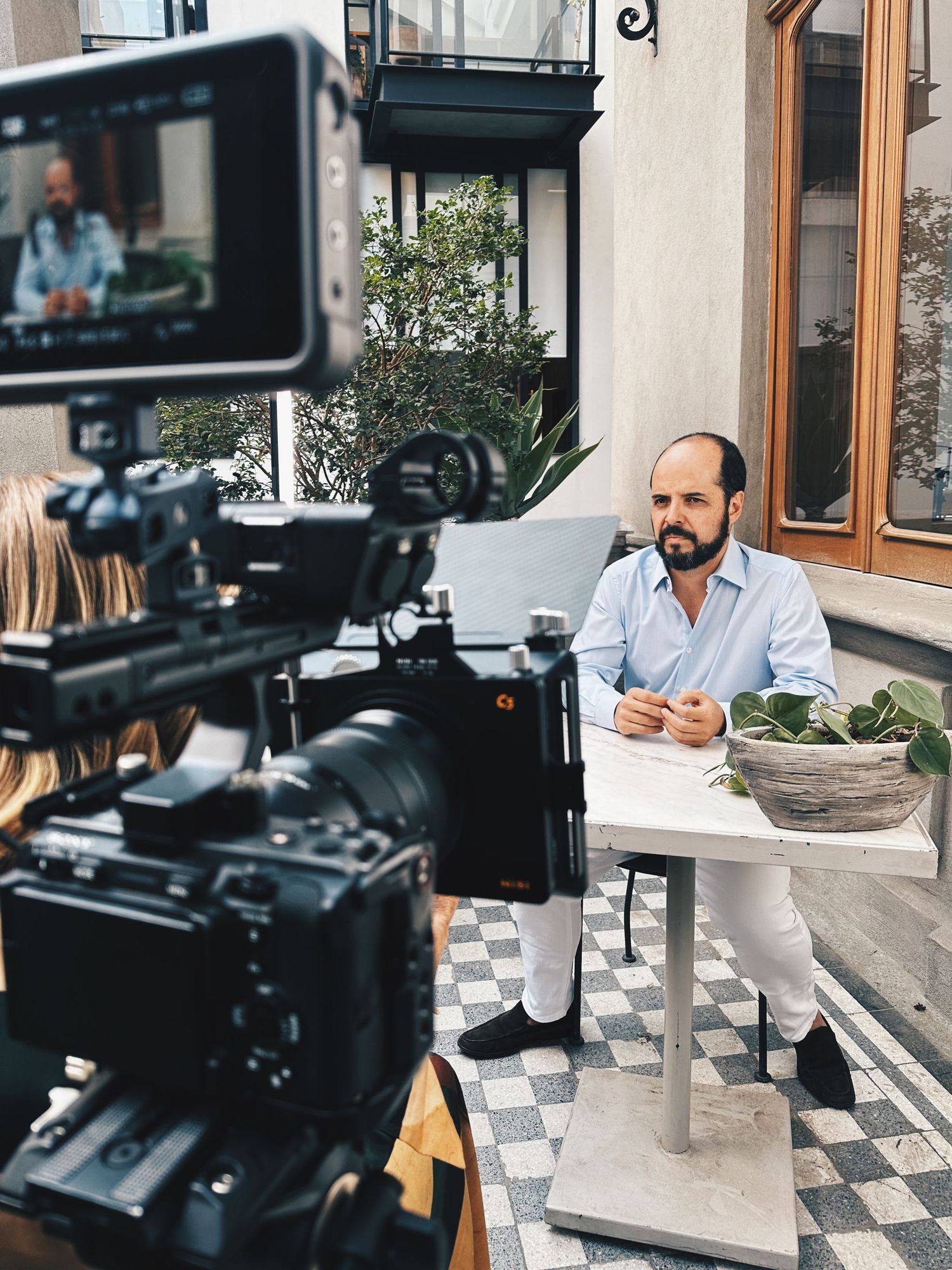Bald bearded man being filmed by multiple cameras during an outdoor interview at a patio table.