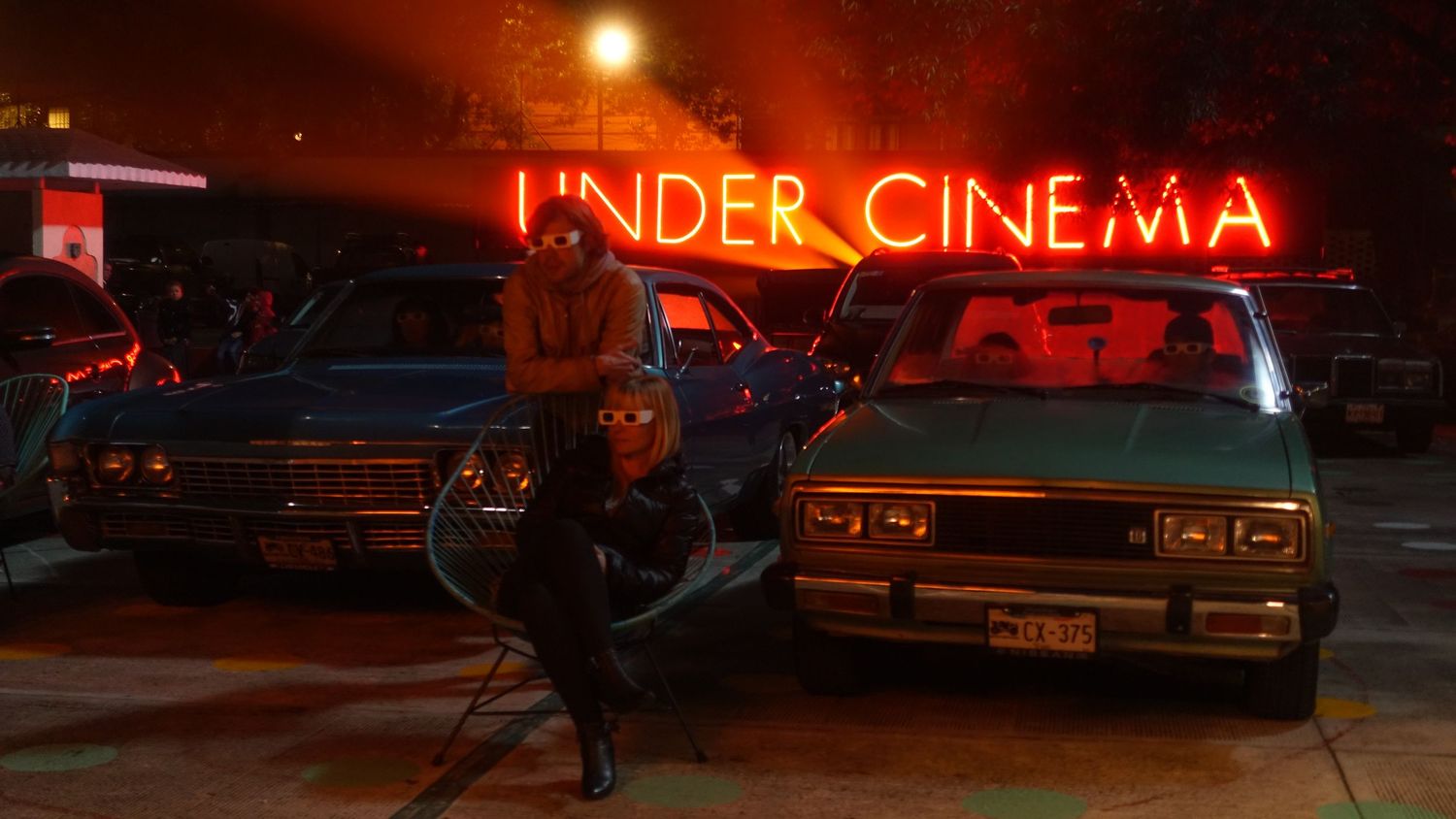 A person poses beside vintage cars under a glowing red neon sign reading Under Cinema at night.