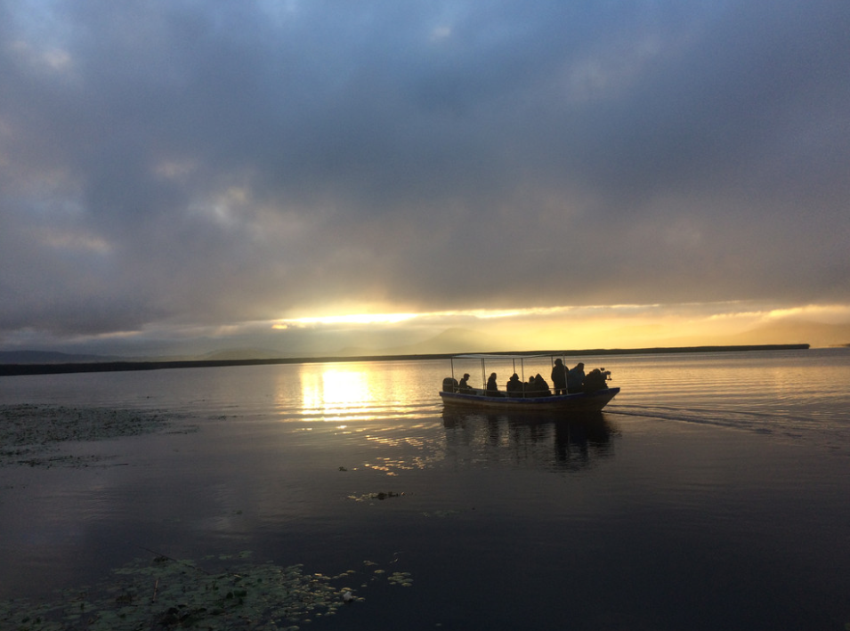 Small boat with passengers silhouetted against a golden sunset over calm lake waters under dramatic clouds.