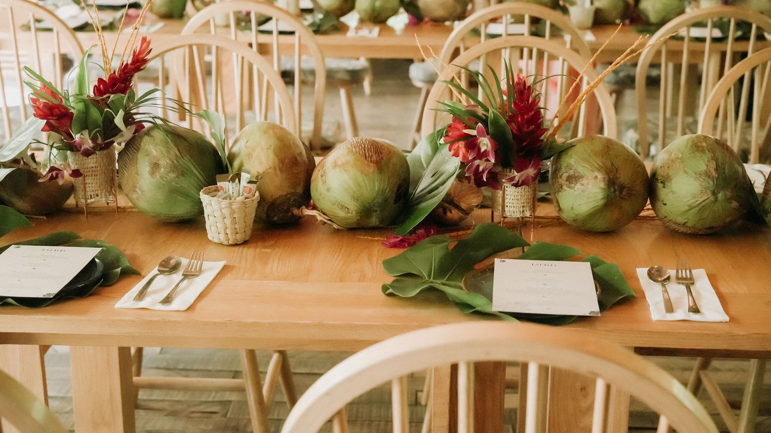 Elegant tropical table setting with coconuts and red floral centerpieces on a wooden dining table.