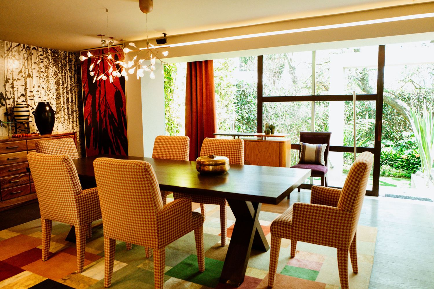 Bright dining room with houndstooth chairs, dark wood table, colorful rug, and garden views.