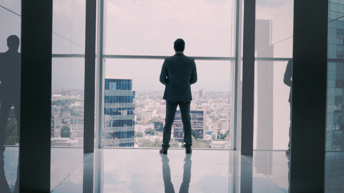 Businessman in suit standing before panoramic floor-to-ceiling windows overlooking modern city skyline.