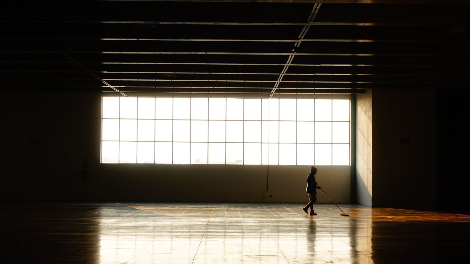 A silhouetted figure walks across a vast, dark gymnasium with a large bright window illuminating the hardwood floor.