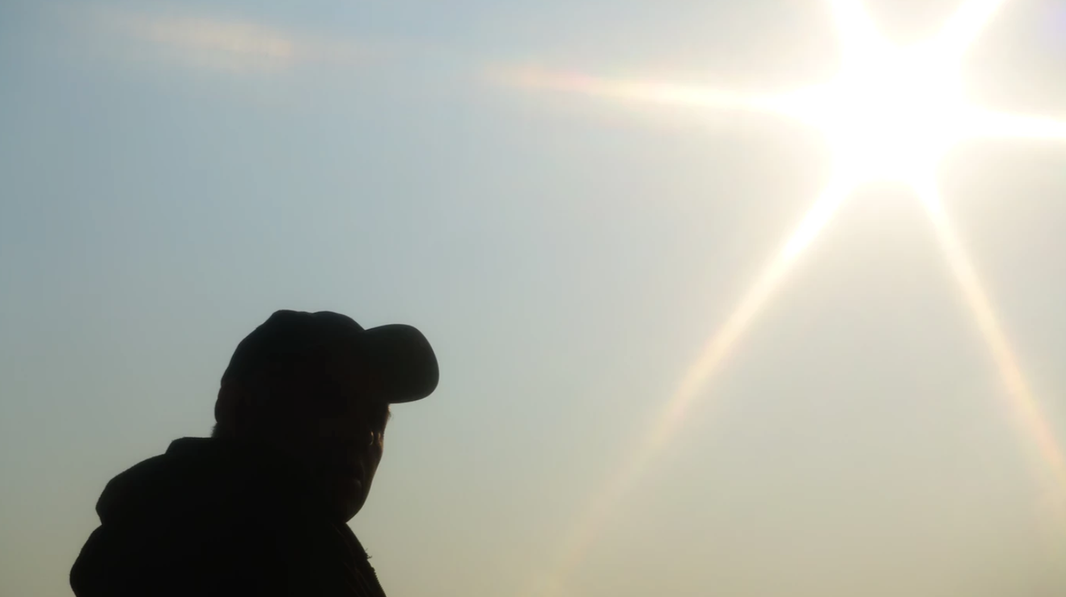 Silhouette of a person wearing a cap against a bright sun and hazy blue sky backdrop.
