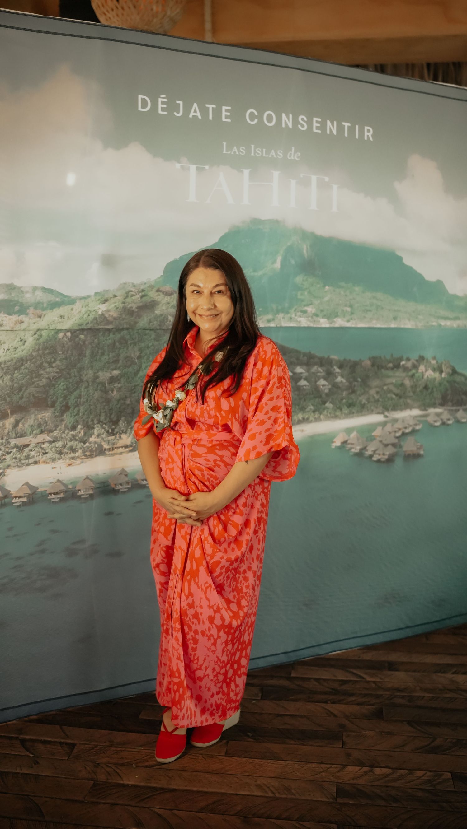 Woman in orange floral dress posing before a Las Islas de Tahiti promotional banner.