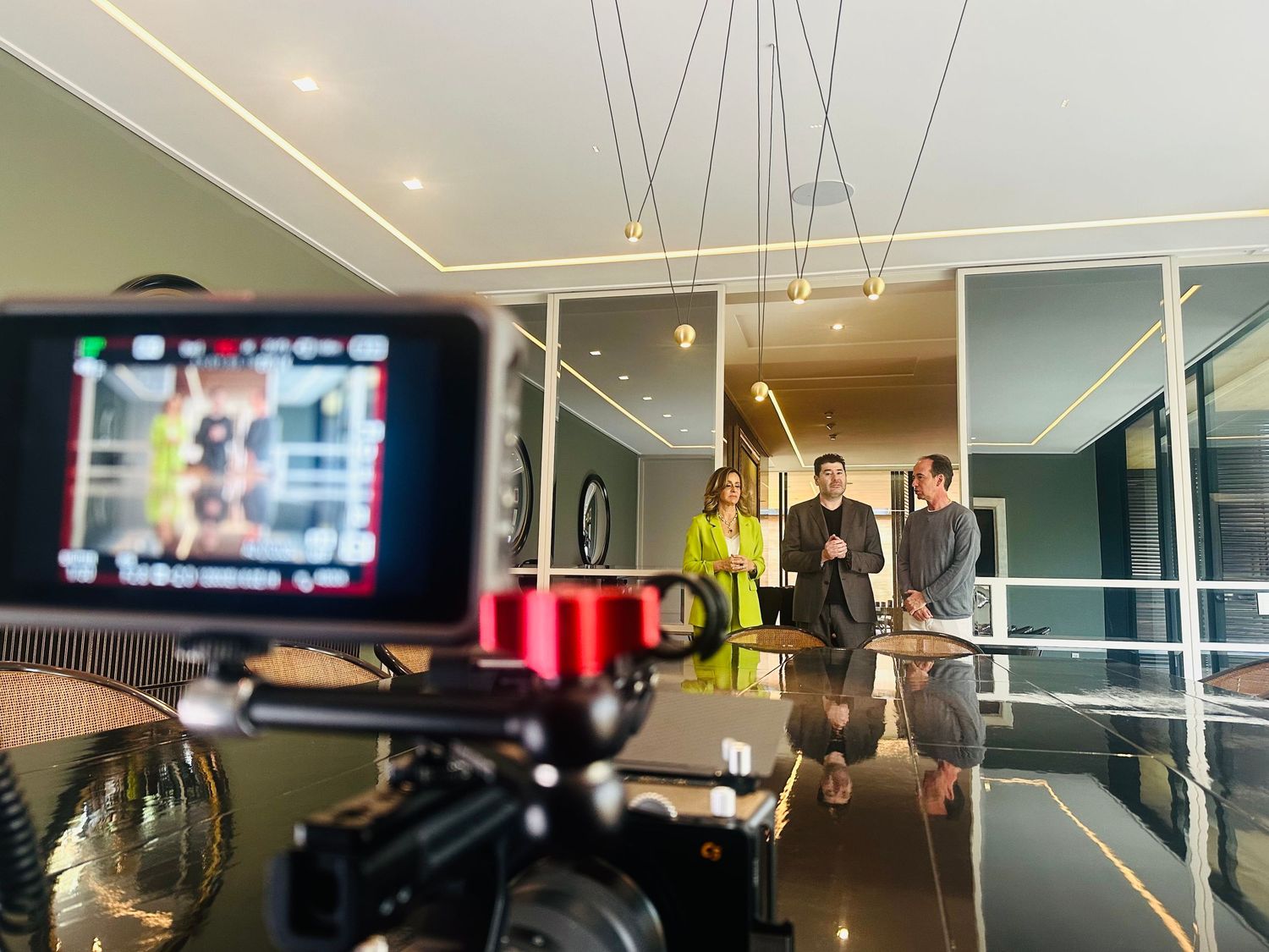 Professional video camera filming three people standing in a modern conference room with pendant lighting.