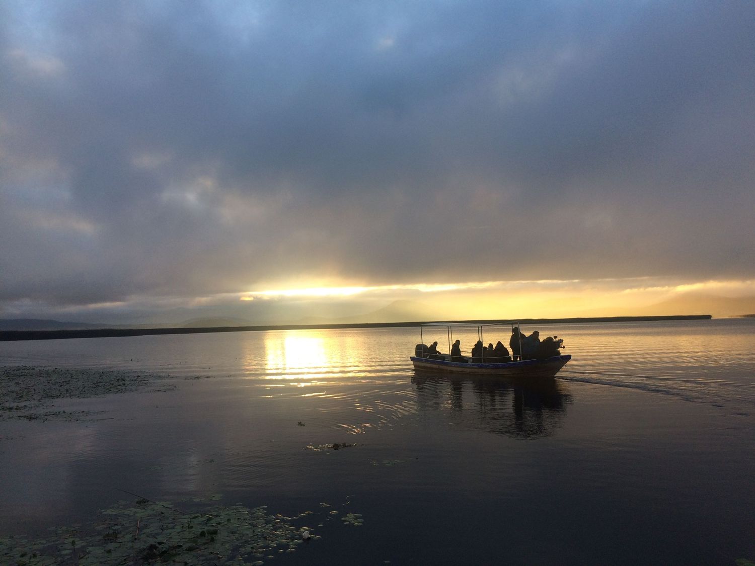 Small boat with passengers crossing a calm lake at golden sunset under dramatic cloudy sky.