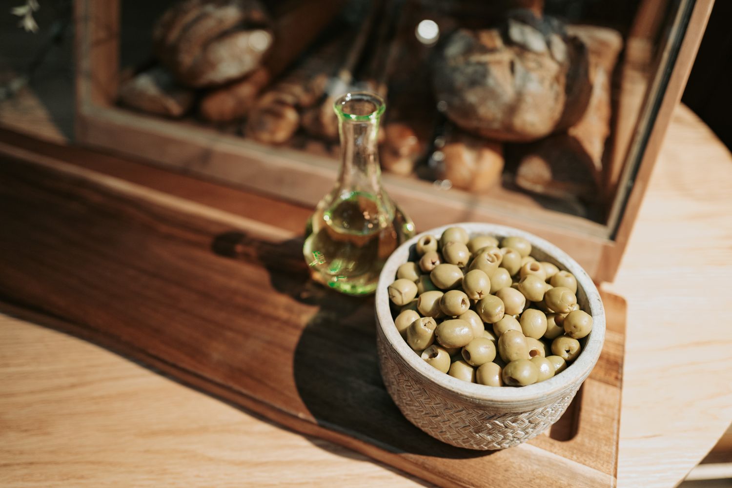 Bowl of green olives beside a glass bottle of olive oil on a wooden shelf with rustic bread loaves in background.