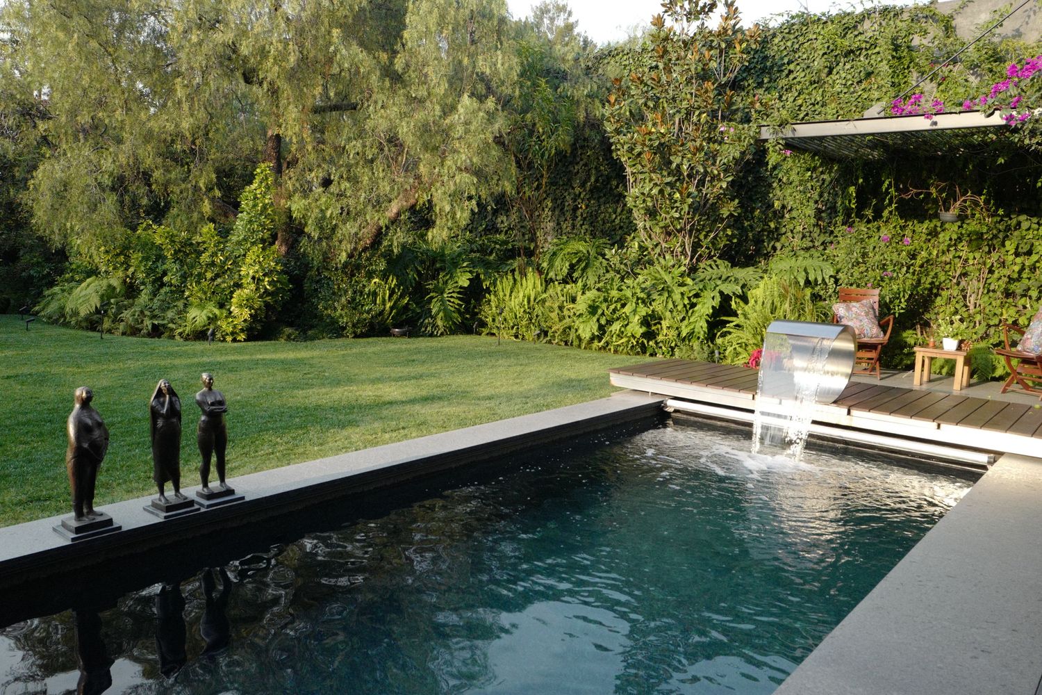 Luxury pool with waterfall feature, bronze sculptures on deck, and lush green garden landscape.
