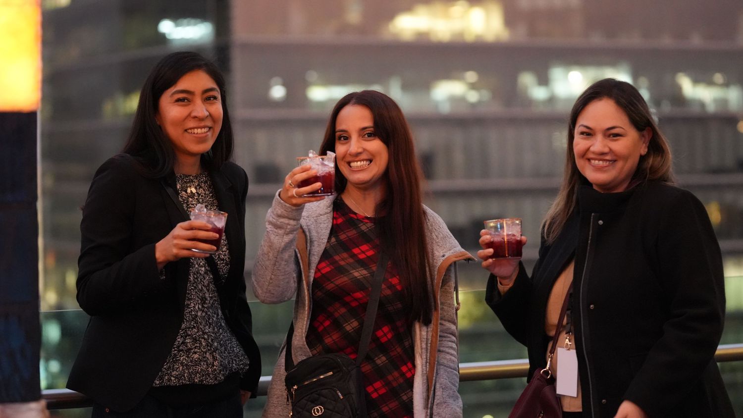 Three women smiling and holding drinks at an outdoor evening networking event.