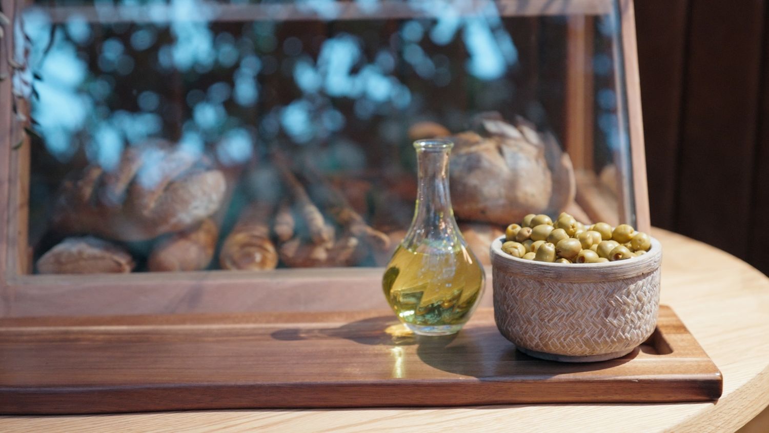 A bottle of olive oil beside a bowl of green olives on a wooden board with bread in the background.