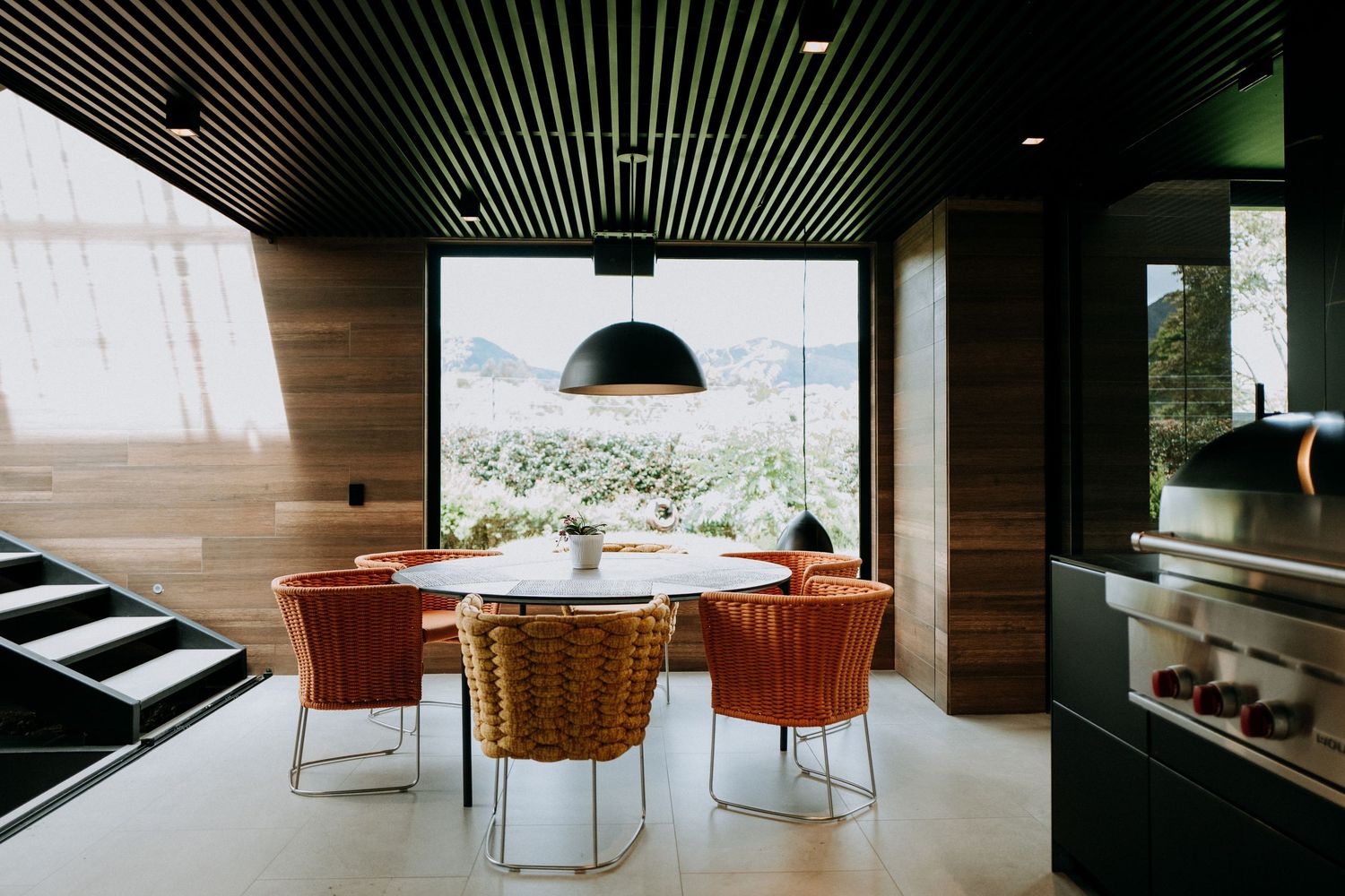 Contemporary dining area with orange chairs, round table, black pendant light, and panoramic mountain view.