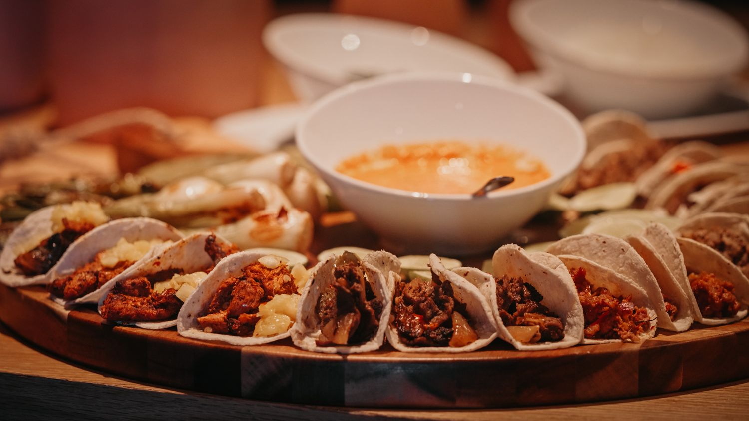 Assorted tacos on a round wooden platter with dipping sauces at a catered event.
