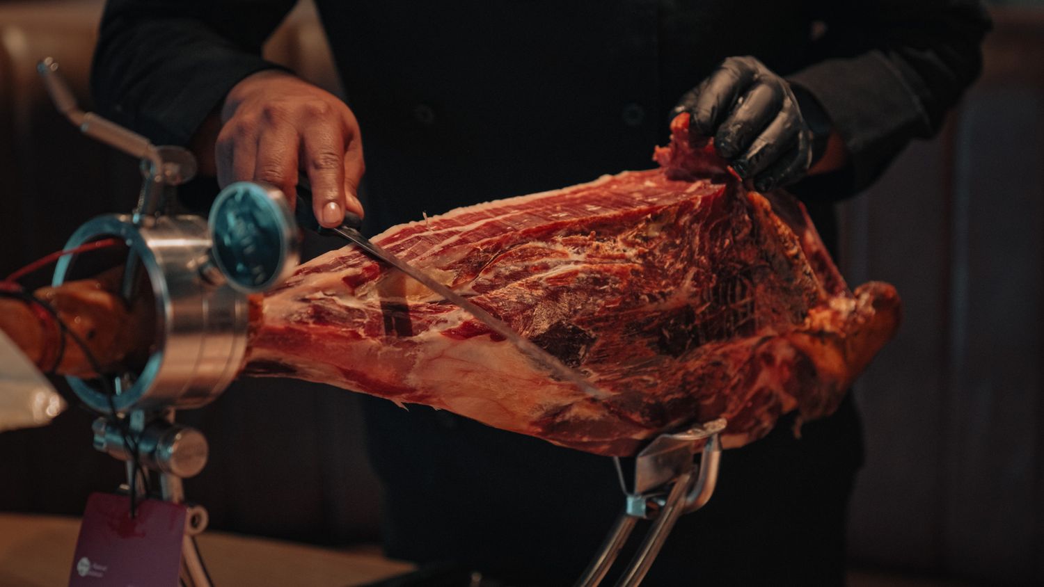 Chef in black gloves expertly carving a whole leg of Iberian ham on a professional jamonero stand.