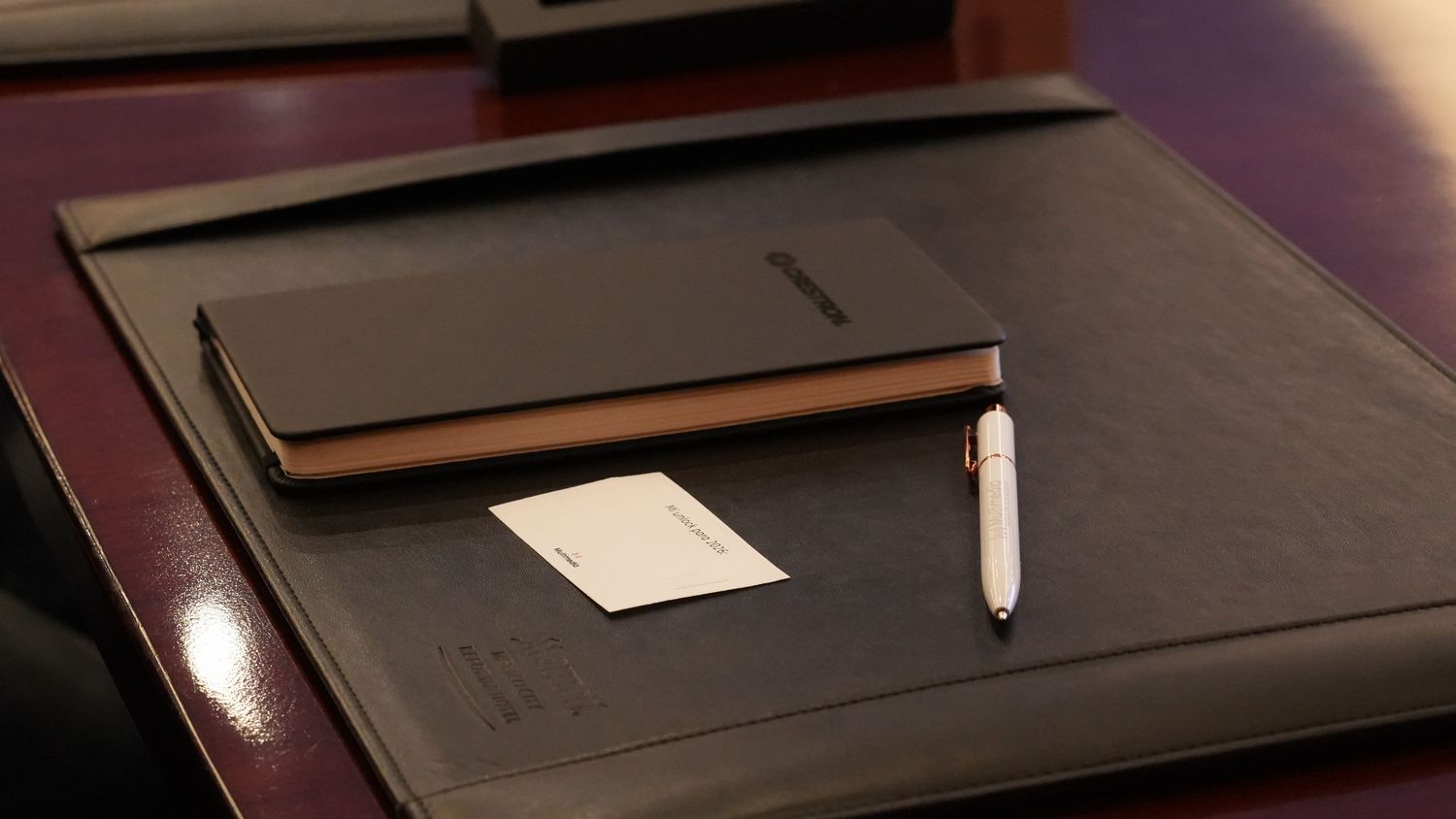 Elegant black leather desk pad with notebook, pen, and business card on a mahogany executive desk.