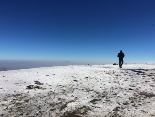 Person walking on snowy mountain summit under clear blue sky.