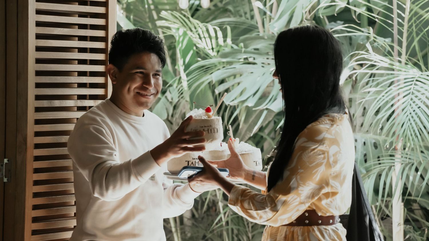 Smiling man and woman in floral dress exchanging tropical coconut drinks in a lush restaurant.
