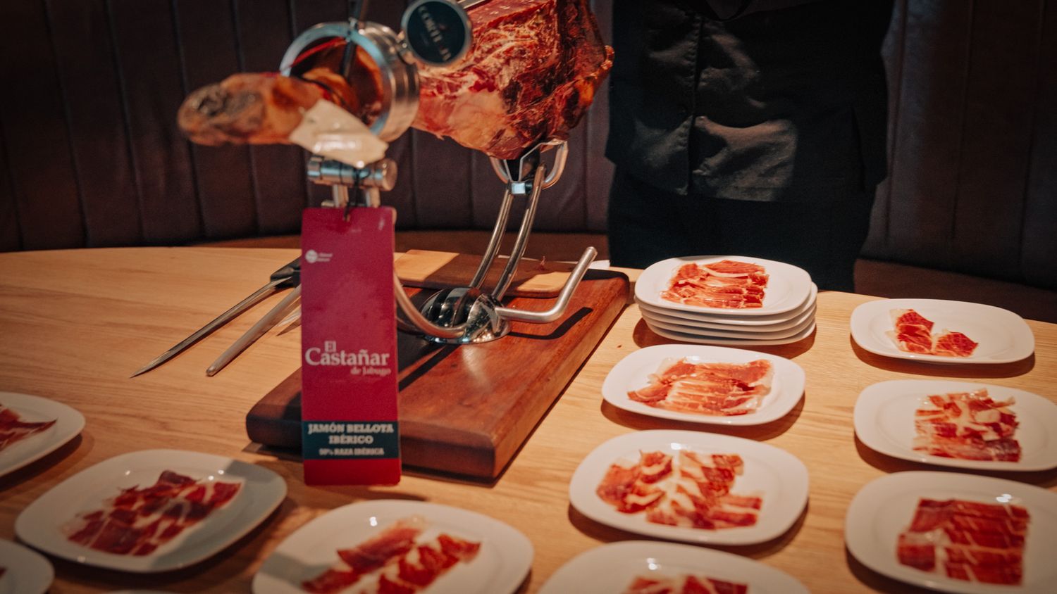 Sliced Iberian ham on white plates displayed beside a jamonero stand at a Cinco Jotas tasting event.