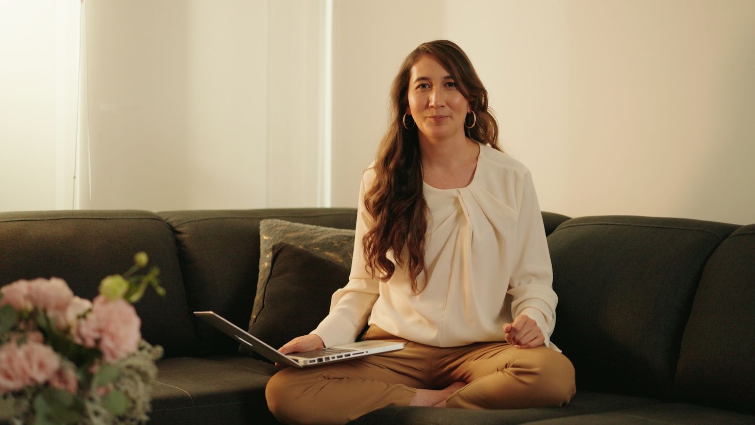 Young woman sitting cross-legged on gray sofa holding laptop in a bright room.