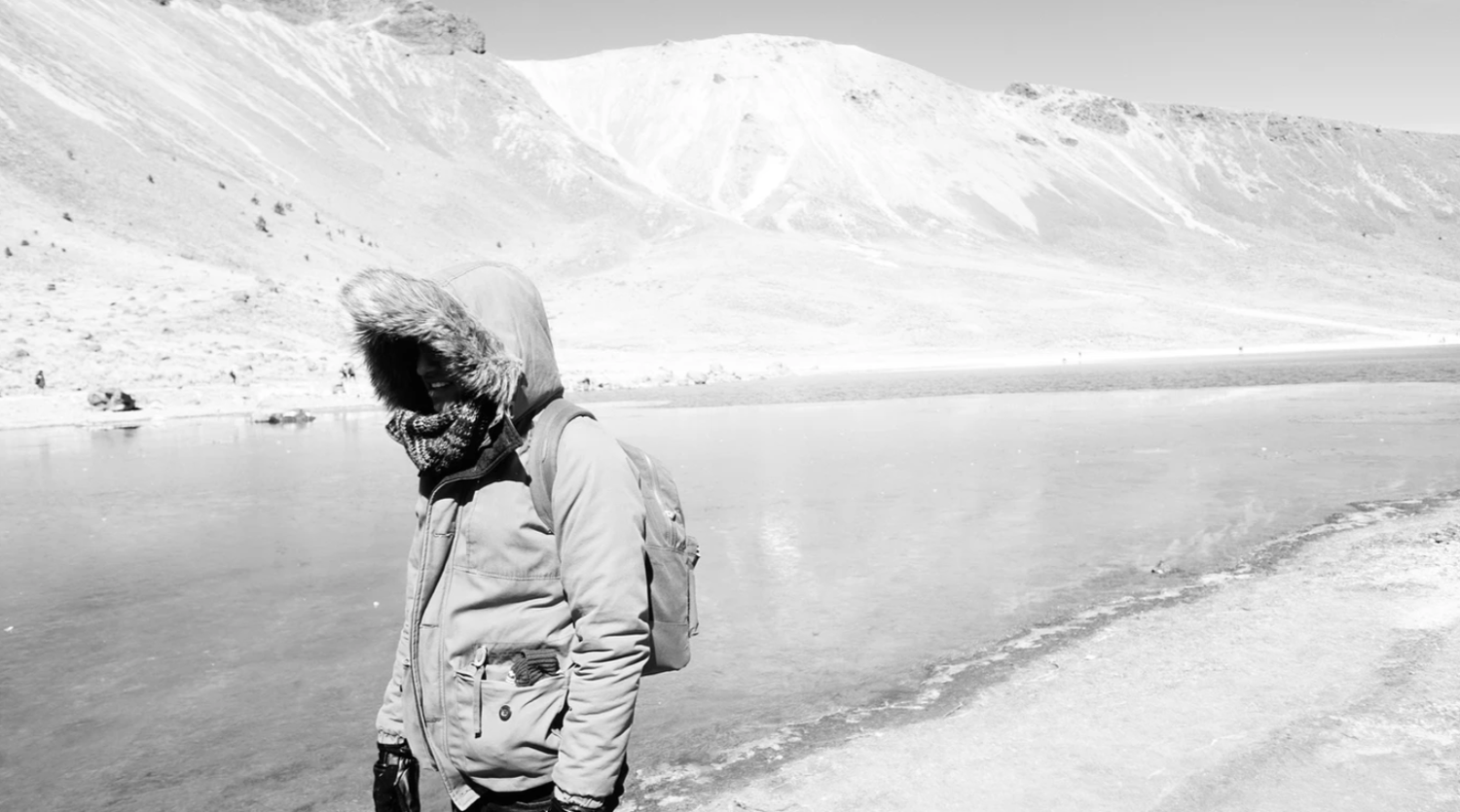Black and white photo of a person in a hooded jacket standing near a frozen lake with snowy mountains in the background.