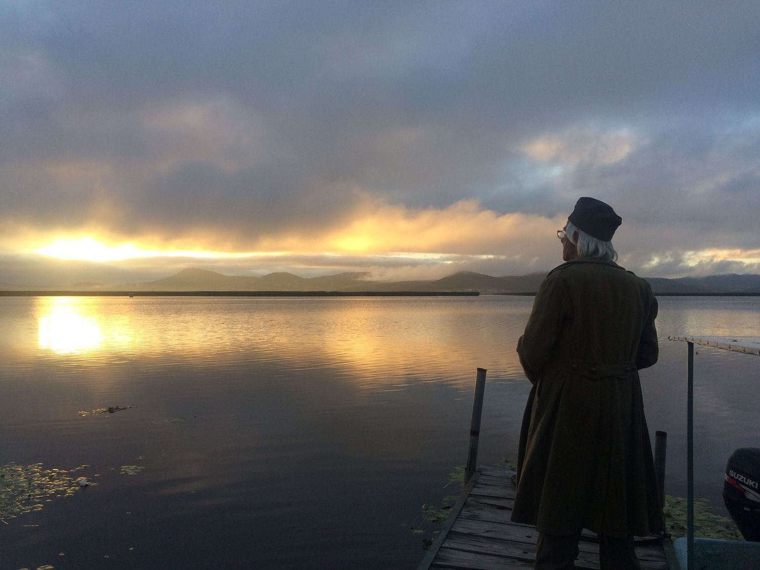 Solitary figure in vintage coat and hat on wooden pier overlooking serene lake at golden sunset.