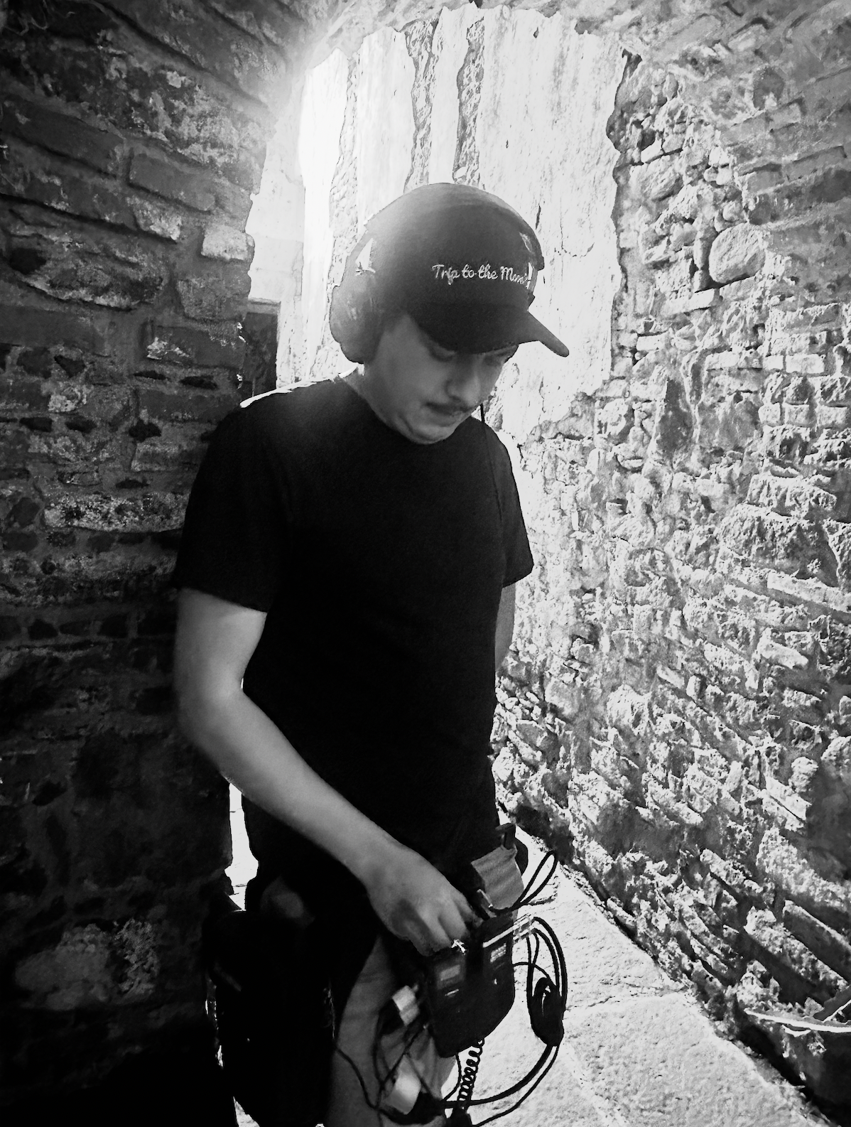 Black and white portrait of young person in cap handling audio equipment against rustic brick wall.