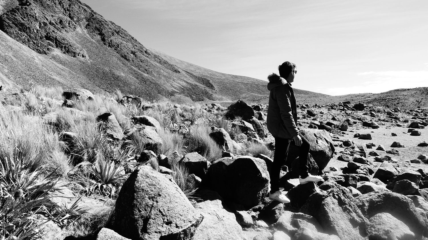 A lone hiker navigates rocky terrain in a dramatic black and white mountain landscape.