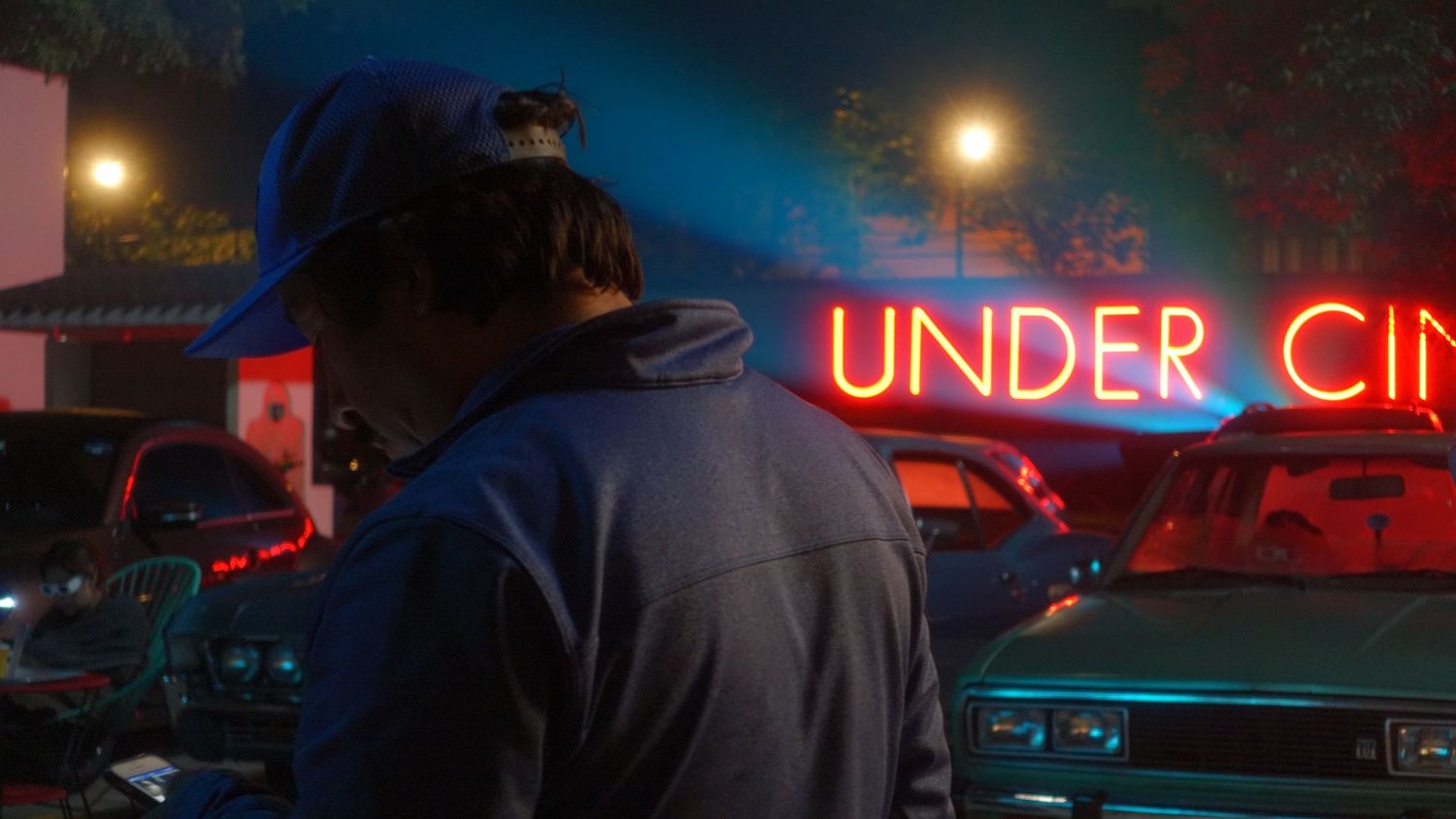 A person stands before a vibrant Under Cinema neon sign surrounded by classic cars at night.