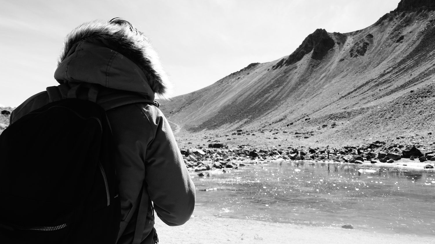 Black and white photo of a person in a hooded jacket overlooking a rocky glacial landscape with mountains.