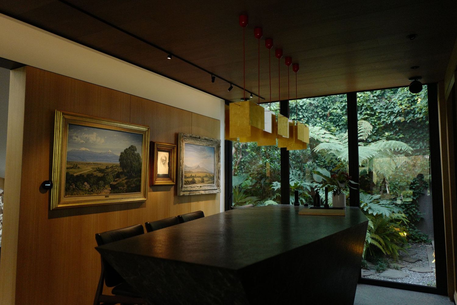 Sophisticated dining room with dark table, gold pendant lights, framed artwork, and garden view.