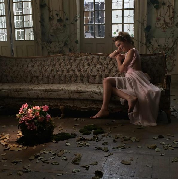 Elegant woman in pink dress sitting on vintage sofa surrounded by rose petals.