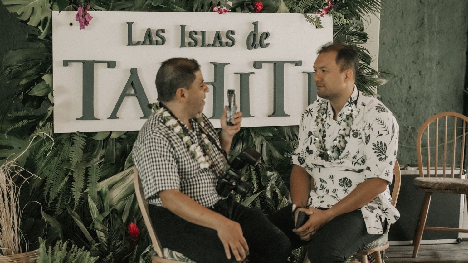 Two men in casual shirts having a conversation in front of a Las Islas de Tahiti sign.