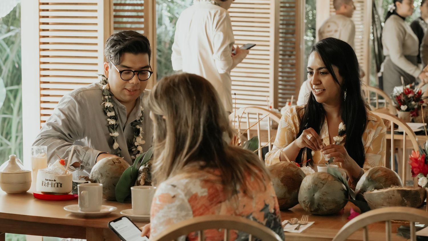 Three people wearing floral leis enjoying conversation at a table with coconuts in tropical restaurant.
