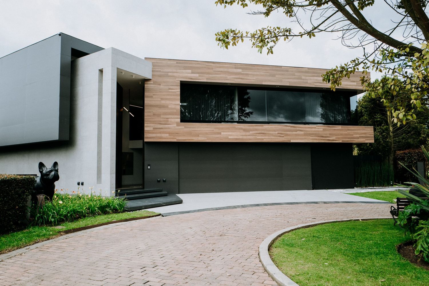 Modern two-story home exterior with wood and concrete facade, dark garage door, and circular driveway.