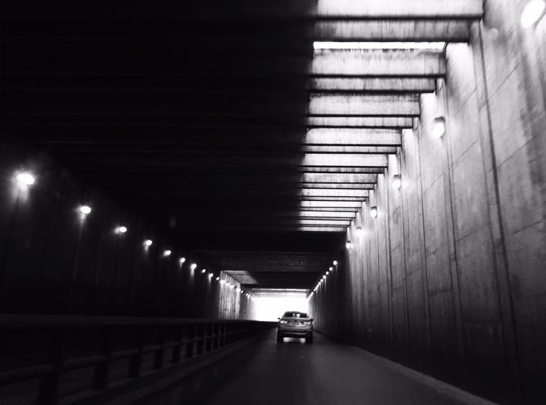 Black and white photo of a car driving through a lit tunnel with concrete walls and ceiling lights.
