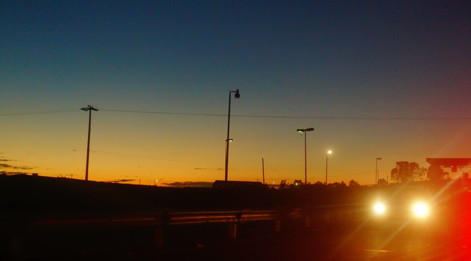 Silhouetted utility poles against a vibrant sunset sky with glowing vehicle headlights illuminating a road at dusk.