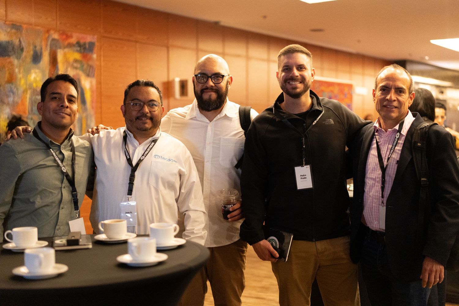 Five professional men smiling together at a networking event with coffee cups on table nearby.